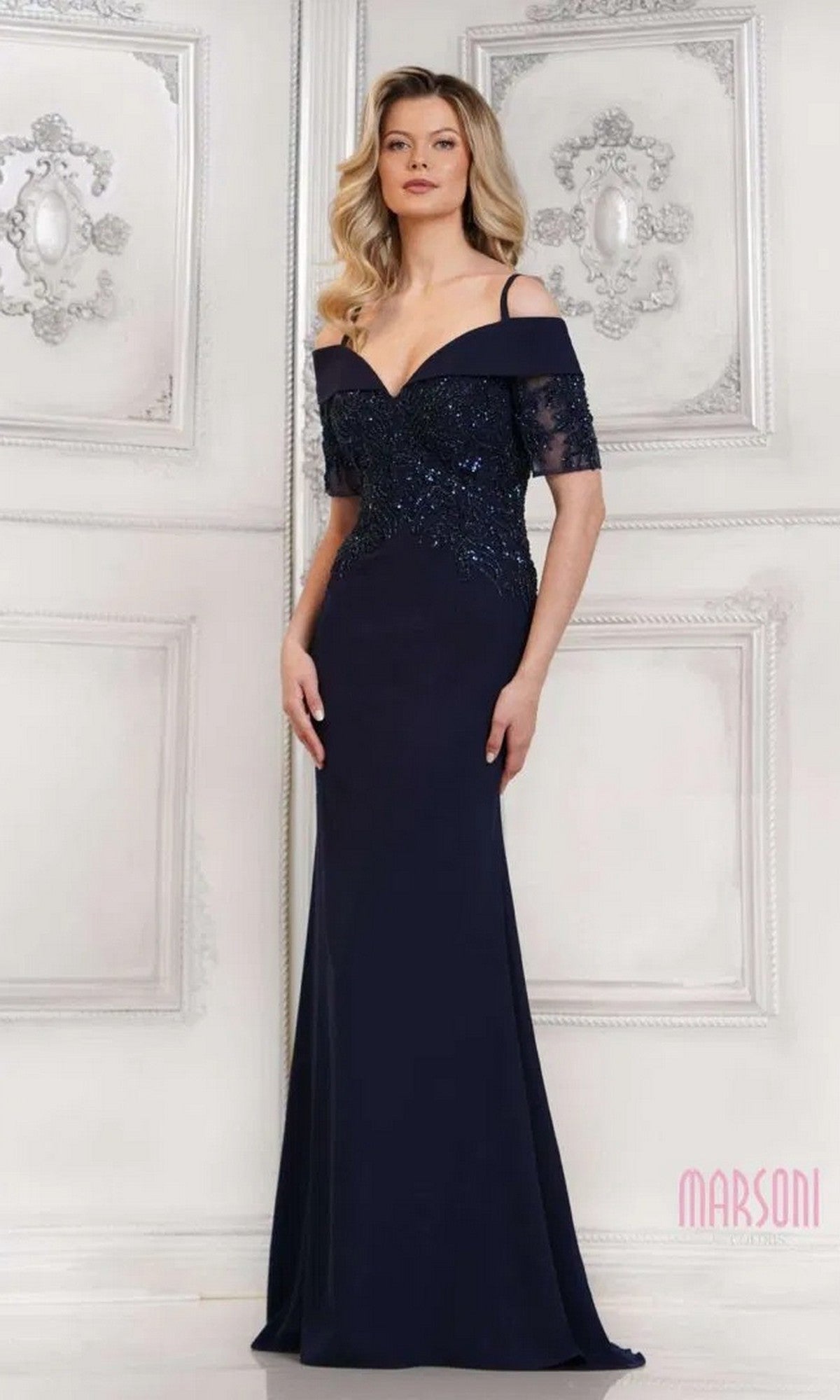 A woman in the Marsoni MV1295 long formal dress in navy, featuring beaded embellishments, stands before an ornate white wall.